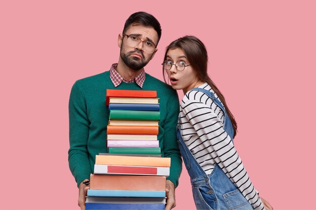 Photo d'un homme mal rasé fatigué avec une expression de pitié, tient une pile de livres, regarde avec mécontentement, sa camarade de classe a regard stupéfié