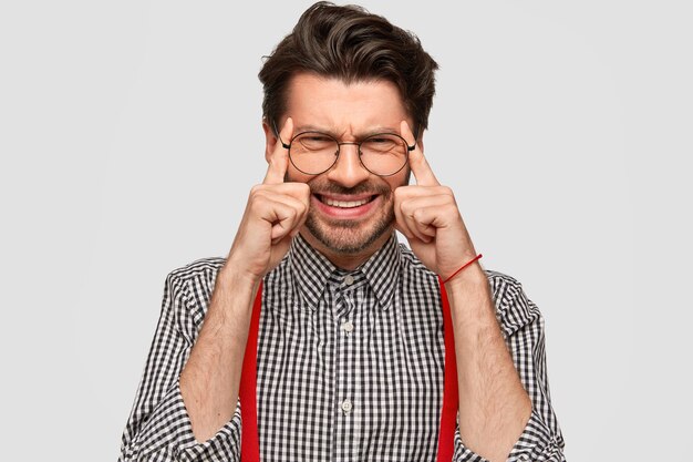 Photo d'un homme mécontent avec chaume et coupe de cheveux à la mode, garde les doigts sur les tempes, essaie de se concentrer sur quelque chose, a mal à la tête, vêtu d'une chemise à carreaux, isolé sur un mur blanc