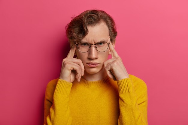 Photo d'un homme mécontent souffre de maux de tête, fronce les sourcils, touche les tempes avec les doigts, essaie de se concentrer, se souvient de quelque chose, porte des lunettes, un pull jaune, isolé sur un mur rose