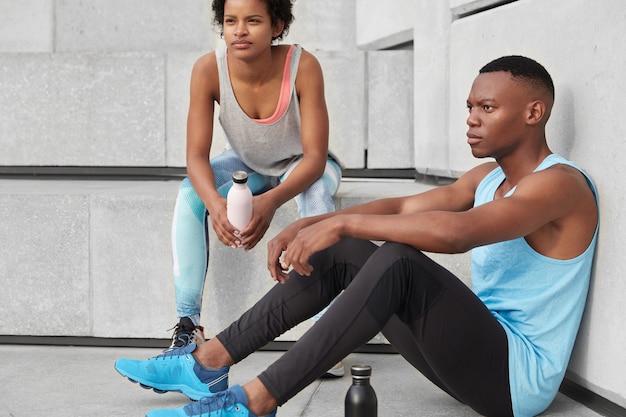 Photo d'un homme motivé à la peau sombre et de sa compagne, posent ensemble dans les escaliers, font une pause après un entraînement cardio en plein air, boivent de l'eau, ont une silhouette athlétique, courent. Concept de mode de vie