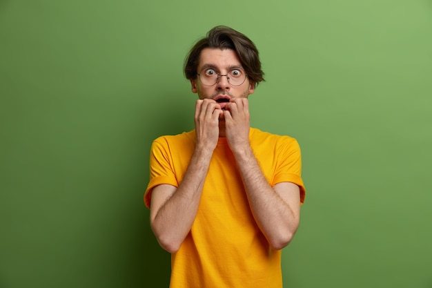 Photo d'un homme nerveux inquiet mord les ongles des doigts et regarde avec une expression effrayée, effrayé par quelque chose de terrifiant, porte des lunettes rondes et un t-shirt jaune, pose contre le mur vert