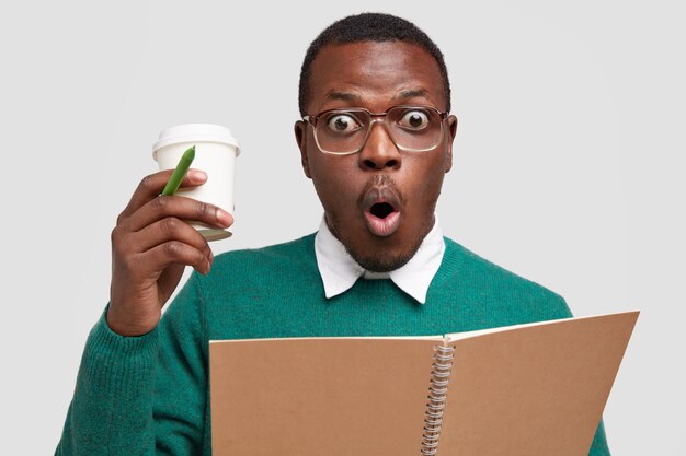 Photo D'un Homme Noir Effrayé Et émotif Regarde Avec Stupeur, Porte Du Café à Emporter, écrit Des Notes Dans Un Cahier, Garde La Bouche Ronde, Habillé Avec Désinvolture