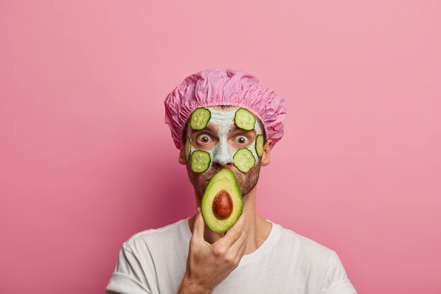 Photo d'un homme surpris couvre la bouche avec la moitié de l'avocat, porte un masque d'argile nettoyante avec des tranches de concombre sur le visage, rafraîchit la peau, réduit les rides