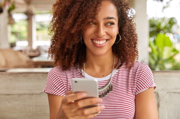 Photo horizontale de contenu heureux Afro American lady détient un cellulaire moderne, reçoit une notification agréable, regarde positivement de côté, vêtu d'un t-shirt décontracté, remarque quelque chose de merveilleux près