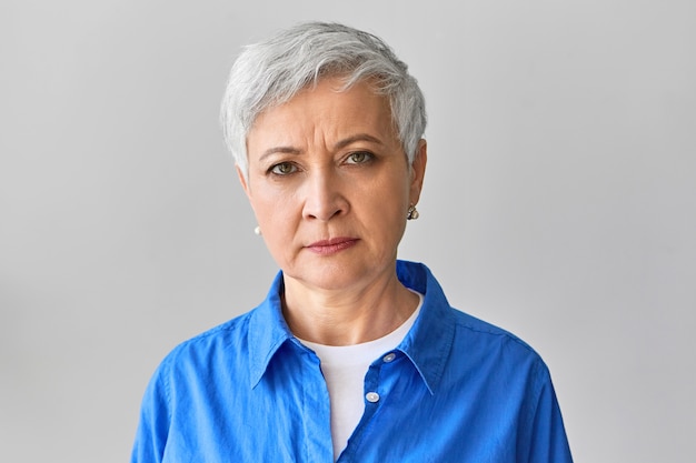 Photo horizontale d'une femme d'affaires mature aux cheveux gris grincheux sérieux en chemise bleue élégante exprimant des émotions négatives, fronçant les sourcils, mécontent de l'employé en retard ou de la date limite manquée