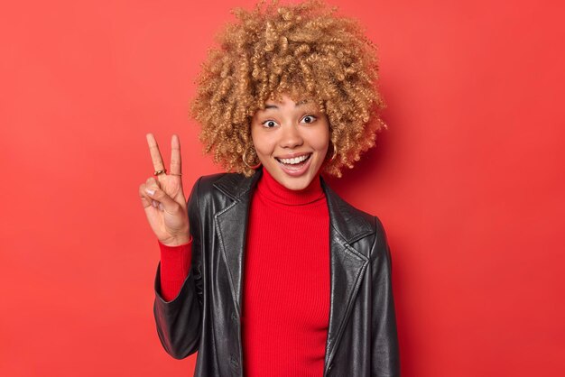Une photo horizontale d'une femme heureuse aux cheveux bouclés fait un signe v montre que le geste de paix a une expression heureuse porte un col roulé et une veste en cuir isolés sur fond rouge vif. Notion de langage corporel