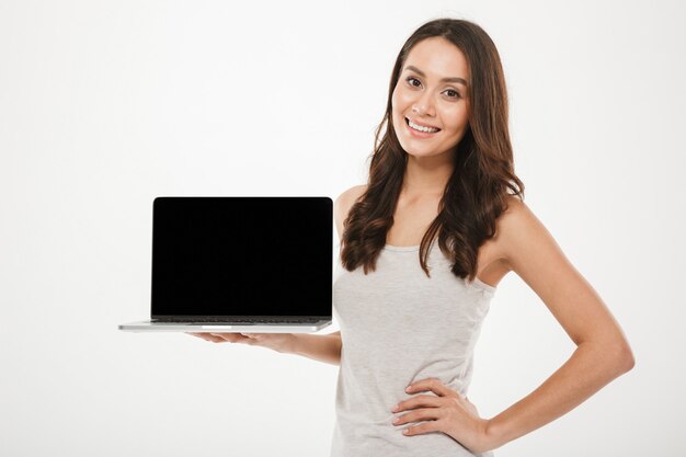 Photo horizontale de femme instruite heureuse souriant et démontrant un écran vide noir d'ordinateur portable en argent tenant sur place, sur mur blanc