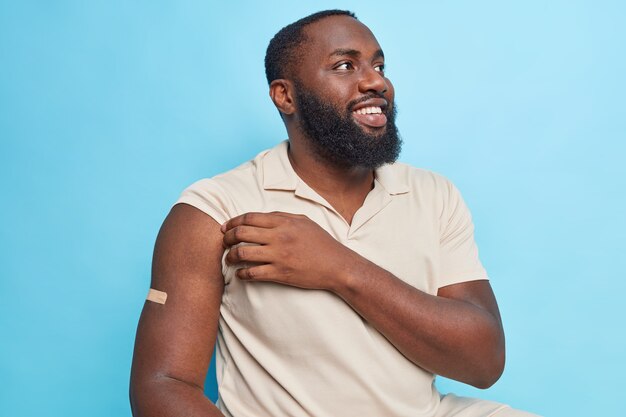 Une photo horizontale d'un homme barbu heureux de se faire vacciner montre le bras après avoir reçu un vaccin engagé dans une campagne de vaccination contre les coronavirus porte des poses de t-shirt décontractées contre le mur bleu