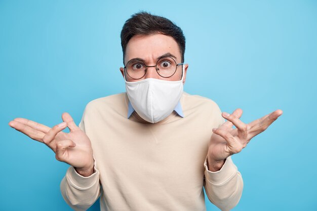 Une photo horizontale d'un homme sérieux porte des lunettes rondes un masque protecteur contre la maladie du coronavirus se propage les mains se sentent confuses ne peuvent pas faire de choix