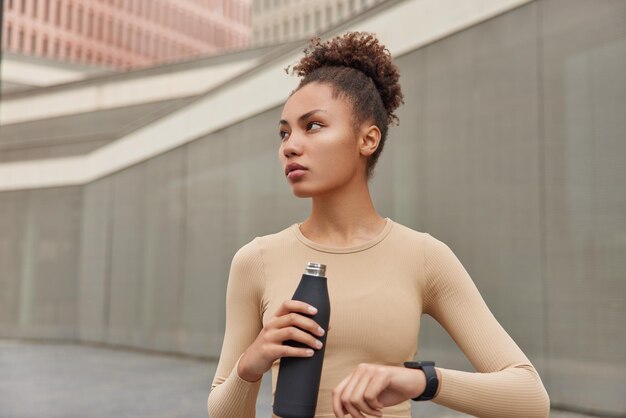 Photo horizontale d'une jeune femme réfléchie aux cheveux bouclés peignés vérifie les résultats de la condition physique sur la smartwatch contient une bouteille d'eau douce pour l'hydratation vêtue de vêtements de sport pose à l'extérieur Mode de vie sain