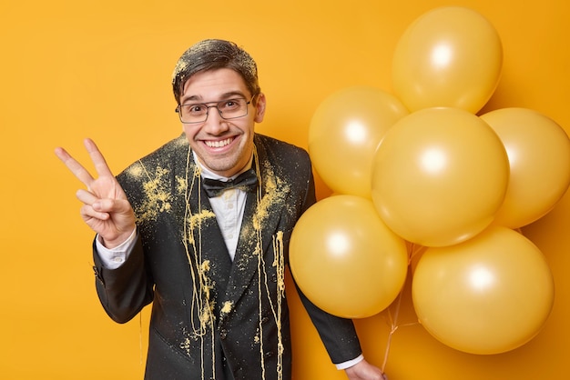 Photo gratuite une photo horizontale d'un jeune homme positif fait un geste de paix célèbre son anniversaire s'amuse lors d'une fête vêtue d'un élégant costume formel tient un tas de ballons gonflés isolés sur fond jaune