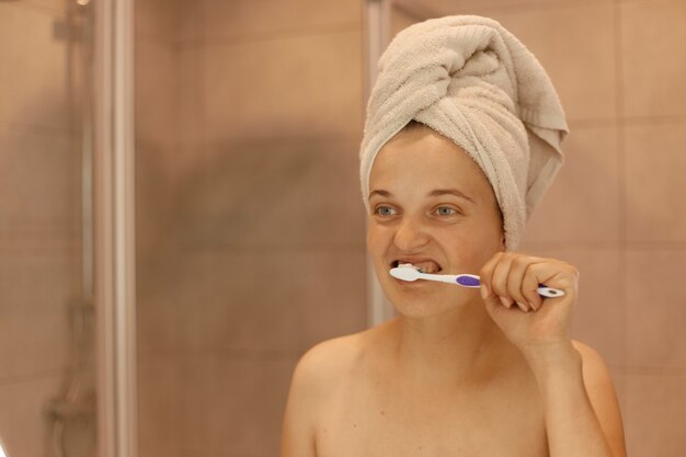 Photo d'intérieur d'une belle jeune fille adulte debout devant le miroir dans la salle de bain et se brossant les dents, faisant des procédures d'hygiène le matin.