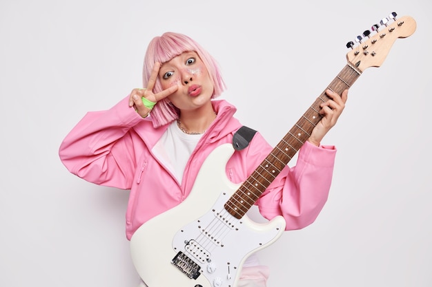 Photo d'intérieur d'une chanteuse de rock n roll aux cheveux roses fait un geste de paix sur les yeux garde les lèvres pliées pose avec une guitare acoustique a une répétition avant le concert