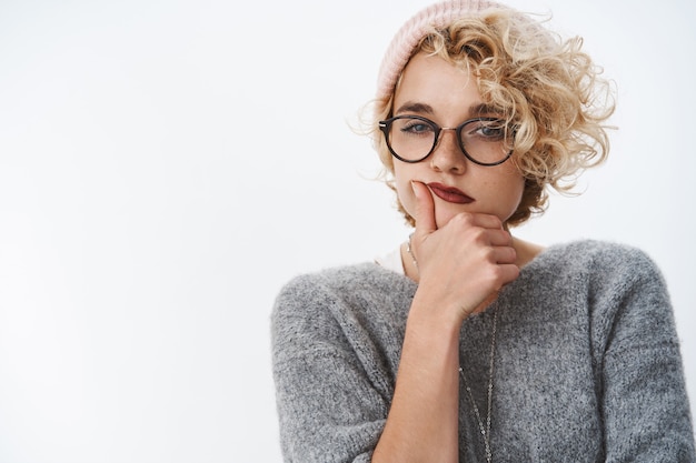 Photo d'intérieur d'une créatrice créative pointilleuse et réfléchie inventant de nouvelles idées portant un bonnet chaud et un pull d'hiver avec des lunettes plissant les yeux en pensant tenant la main sur le menton sur un mur blanc