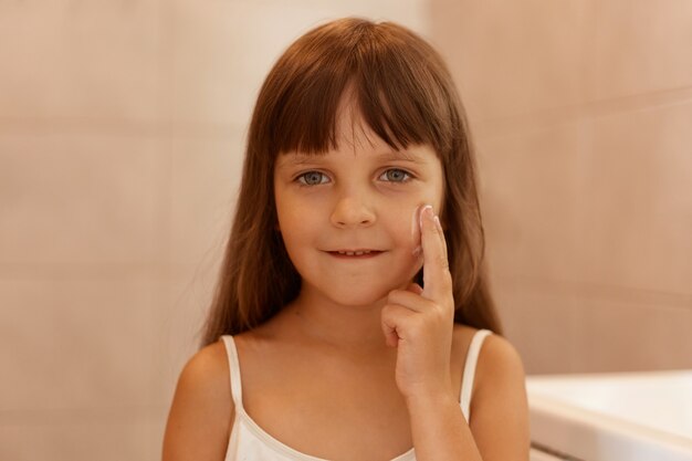 Photo d'intérieur d'une enfant de sexe féminin avec de la crème pour le visage sur la joue, pointant le doigt sur son visage, regardant directement la caméra, portant un t-shirt blanc sans manches, faisant seule des procédures de beauté.
