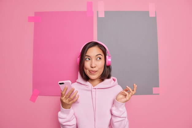 Photo d'intérieur d'une femme asiatique rêveuse léchant les lèvres imagine quelque chose tout en écoutant sa musique préférée utilise les technologies modernes passe du temps libre à profiter de nouvelles poses de plylist contre un espace de copie vierge