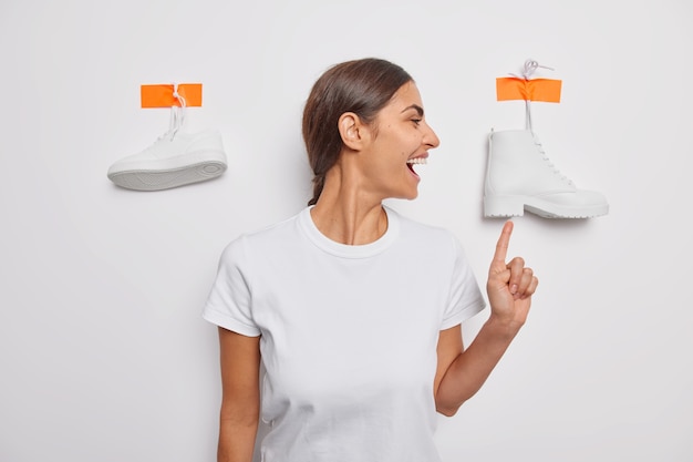 Une photo d'intérieur d'une femme brune joyeuse montre des chaussures pour porter des chaussures blanches comme neige après avoir marché vêtu de poses de t-shirt décontractées seules