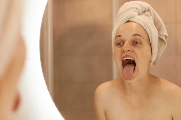 Photo d'intérieur d'une femme ensoleillée posant dans la salle de bain après avoir pris une douche, regardant son reflet dans un miroir avec la langue sortie, regarde sa langue, procédures hygiéniques.