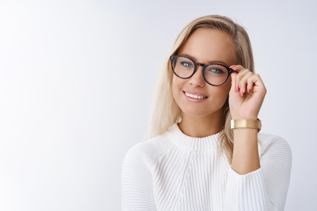 Photo gratuite photo d'intérieur d'une femme entrepreneur intelligente et prospère partageant les moyens de réussir en touchant des montures vérifiant des lunettes souriantes ravies et accomplies en regardant la caméra avec assurance contre un mur blanc.