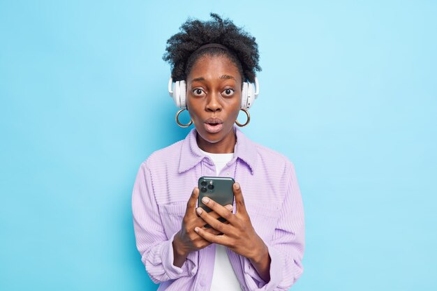 Photo d'intérieur d'une femme à la peau foncée surprise qui tient un téléphone portable et écoute de la musique via des écouteurs