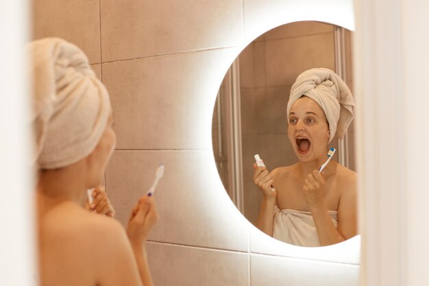 Photo d'intérieur d'une femme avec une serviette blanche sur la tête tenant du dentifrice et une brosse à dents dans les mains, regardant son reflet dans le miroir avec la bouche largement ouverte.