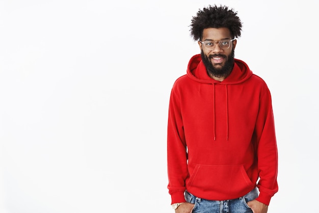 Photo gratuite photo d'intérieur d'un jeune afro-américain audacieux et beau nomade numérique mâle barbu dans des verres avec une coiffure afro portant un sweat à capuche rouge et cool