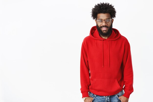 Photo d'intérieur d'un jeune afro-américain audacieux et beau nomade numérique mâle barbu dans des verres avec une coiffure afro portant un sweat à capuche rouge et cool