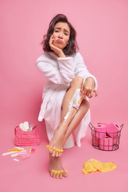 Photo gratuite photo d'intérieur d'une jeune femme asiatique malheureuse aux cheveux noirs se rasant les jambes avec une expression mécontente étant pressée tard pour le rendez-vous, la pédicure porte un peignoir blanc confortable pose sur la cuvette des toilettes