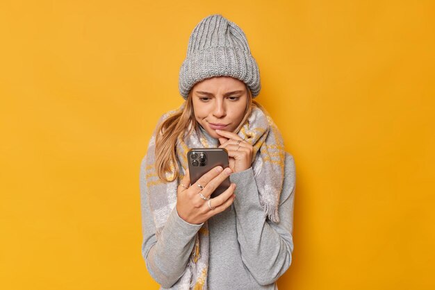 Une photo d'intérieur d'une jeune femme attentive concentrée avec une expression sérieuse à l'écran du smartphone reçoit un message dérangeant regarde la vidéo avec attention, porte un pull et une écharpe tricotés chauds.