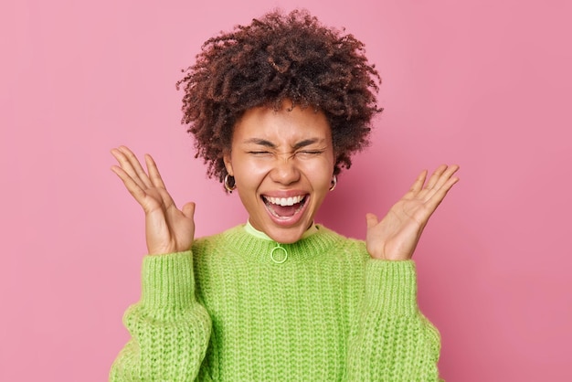 Photo gratuite une photo d'intérieur d'une jeune femme émotionnelle aux cheveux bouclés crie fort et garde les paumes levées la bouche ouverte devenant folle vêtue de poses de pull tricoté vert décontracté sur fond rose se sent excité.