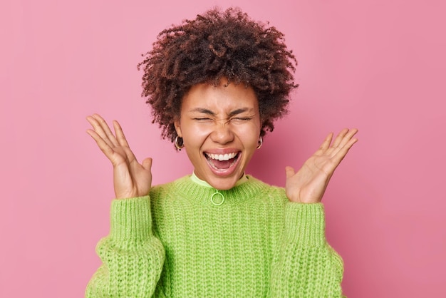 Une photo d'intérieur d'une jeune femme émotionnelle aux cheveux bouclés crie fort et garde les paumes levées la bouche ouverte devenant folle vêtue de poses de pull tricoté vert décontracté sur fond rose se sent excité.
