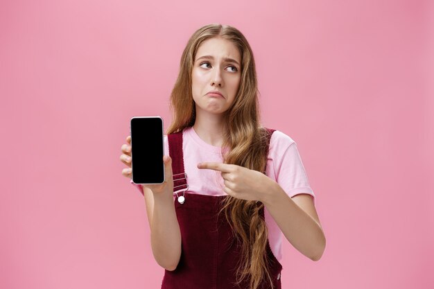 Photo d'intérieur d'une jolie jeune fille sombre, mécontente et déçue, tenant un smartphone pointant vers cel...