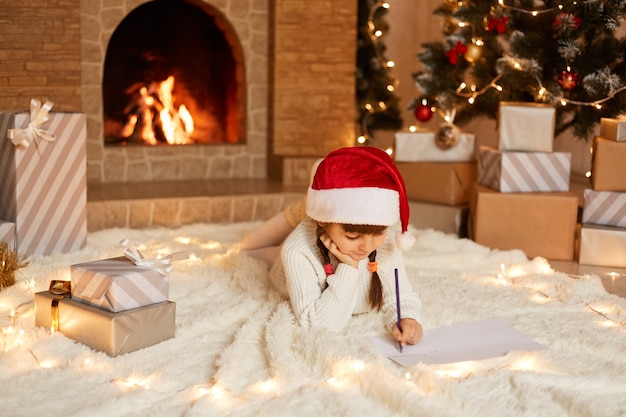 Photo d'intérieur d'une petite fille mignonne portant un pull blanc et un chapeau rouge, allongée sur le sol sur un tapis moelleux dans une pièce décorée de fête, écrivant une lettre au père Noël.