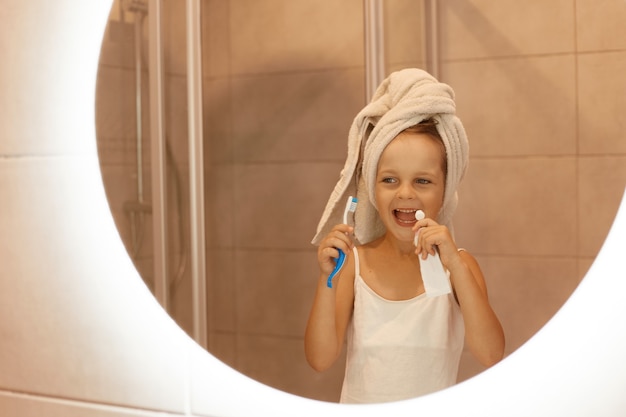 Photo d'intérieur d'une petite fille se brossant les dents dans la salle de bain, regardant son reflet dans le miroir avec une expression faciale excitée, portant un t-shirt blanc et enroulant ses cheveux dans une serviette.