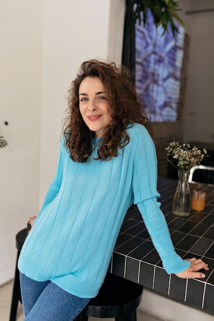 Photo Intérieure D'une Belle Fille Avec Des Boucles En Pull Bleu Tendre Sourit à La Caméra Et Se Repose Au Café