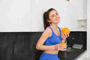 Photo gratuite une photo intérieure d'une femme après l'entraînement debout dans la cuisine avec du jus frais et une orange à boire