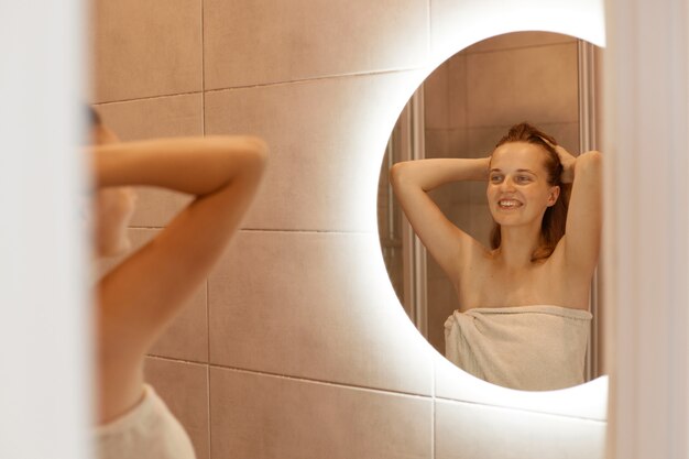 Photo intérieure d'une femme souriante regardant son reflet dans un miroir avec un charmant sourire heureux, debout les bras levés, une femme étant enveloppée dans une serviette blanche.