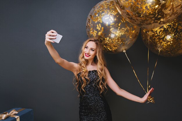 Photo intérieure d'une fille d'anniversaire blonde romantique faisant selfie. Femme caucasienne frisée souriante se détendre à la fête avec des ballons et des cadeaux.