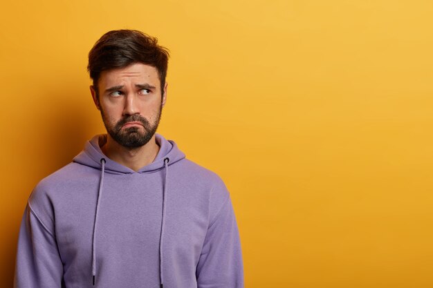 Une photo intérieure d'un homme sombre et insatisfait se sent déprimé et solitaire, désespéré, réfléchit à certains problèmes, fronce les sourcils, habillé avec désinvolture, pose contre le mur jaune, espace libre de côté