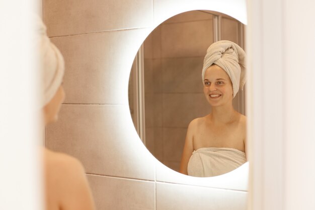 Photo intérieure d'une jeune femme adulte séduisante regardant le reflet dans le miroir après la douche et souriante, enveloppée dans une serviette blanche, exprimant des émotions positives.