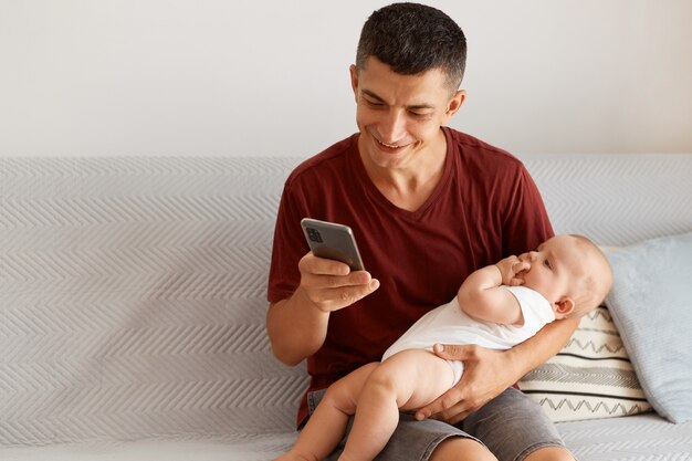 Photo intérieure d'un jeune homme adulte portant un t-shirt marron de style décontracté, assis sur un canapé gris dans la chambre, tenant un bébé en bas âge, utilisant un téléphone intelligent, souriant joyeusement.