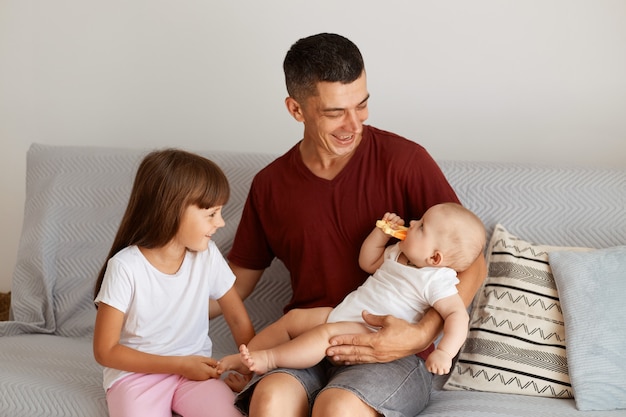 Photo intérieure d'un père heureux portant un t-shirt de style décontracté bordeaux assis sur un canapé avec ses filles, regardant avec amour et douceur son bébé, riant, appréciant de passer du temps ensemble.