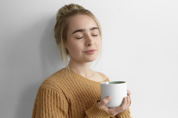 Photo isolée de la belle jeune femme portant un pull chaud confortable fermant les yeux avec plaisir, appréciant le café frais chaud, tenant une grande tasse