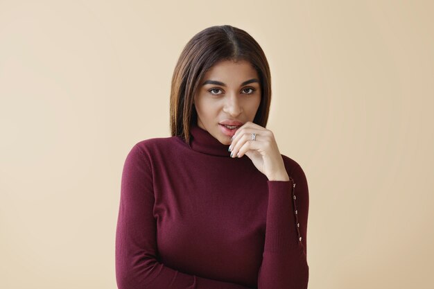 Photo isolée d'élégante magnifique jeune femme à la peau sombre portant un pull à col roulé violet à la mode posant, regardant avec un regard ludique mystérieux, touchant ses lèvres en peluche