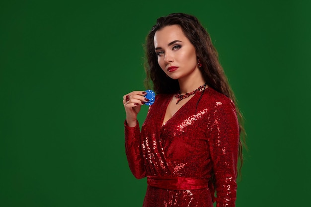Photo de jeune belle femme brune chanceuse sexy dans une robe de soirée rouge brillante tient quelques jetons de poker dans ses mains. fond vert, chromakey, industrie du jeu