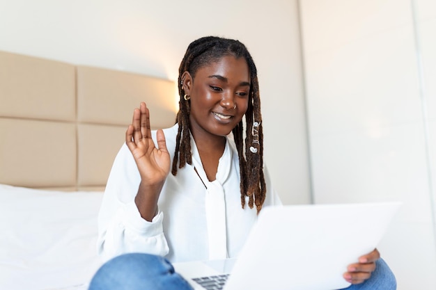 Photo gratuite photo d'une jeune femme africaine souriante qui passe un appel vidéo la nuit assise sur un lit dans sa chambre