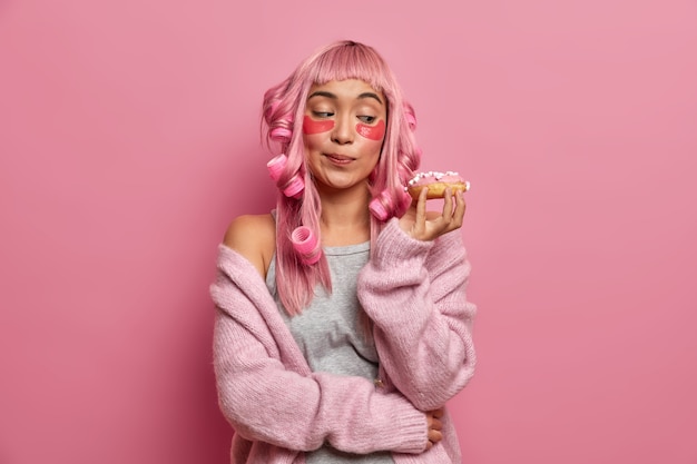 Photo de jeune femme asiatique regarde beignet appétissant, fait les cheveux avec des rouleaux, applique des tampons de collagène, habillé en pull chaud