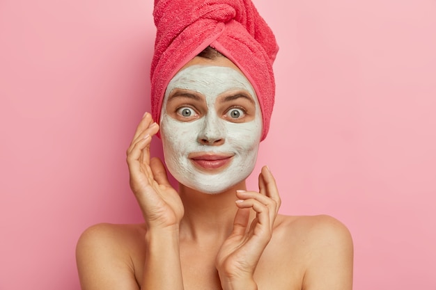 Photo d'une jeune femme attaractive applique un masque nutritif pour les soins du visage, veut avoir une peau fraîche et propre, porte une serviette rose sur la tête