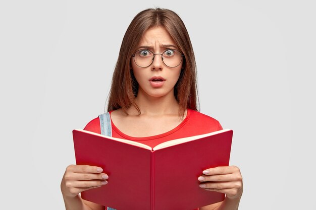 Photo d'une jeune femme caucasienne émotive stupéfaite regarde avec stupéfaction, tient le livre rouge, doit apprendre beaucoup pour la prochaine leçon, porte des lunettes rondes, isolé sur un mur blanc