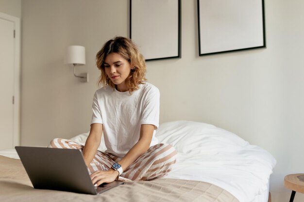 Photo d'une jeune femme avec une coiffure ondulée portant un pyjama blanc est assise dans le lit le matin et travaille sur un ordinateur portable au soleil dans un appartement moderne et élégant